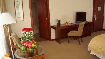 Una habitación de hotel con una cama, un escritorio con computadora, una silla, una mesita con flores y una lámpara.