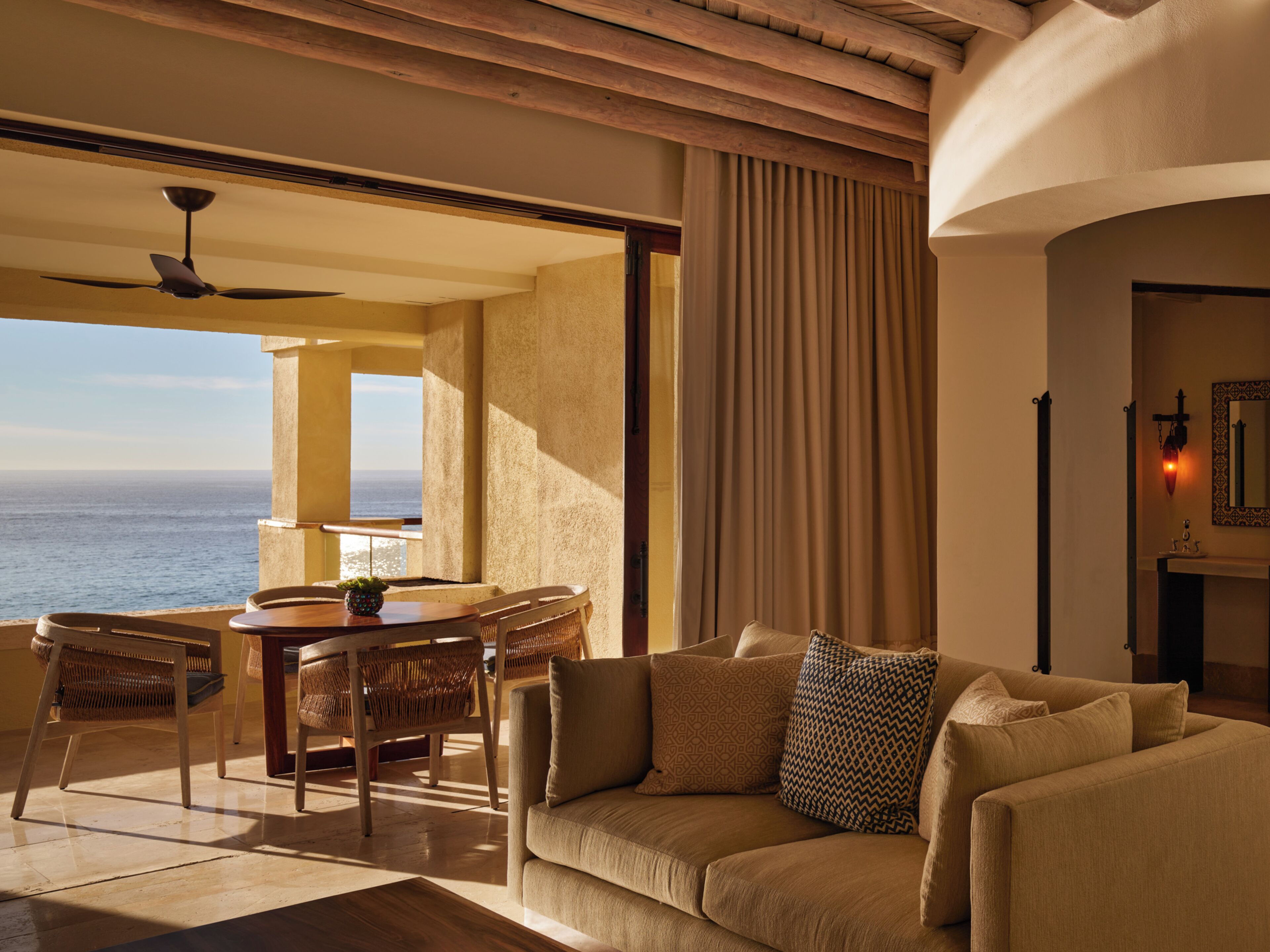 A room with a sofa, a dining table with chairs, a view of the ocean, and a ceiling fan.