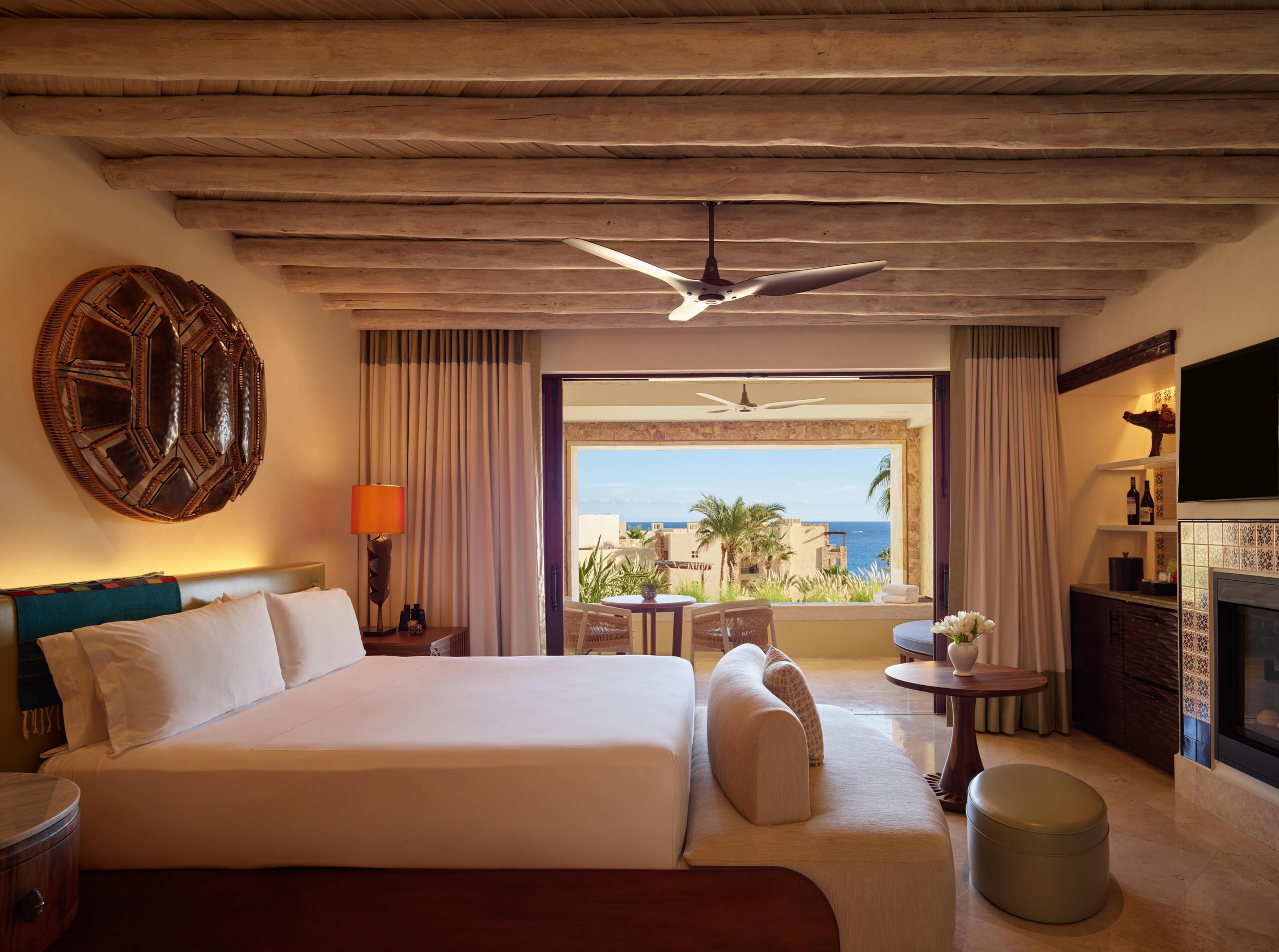 A hotel room with a large bed, a ceiling fan, a view of the sea, a dining area, and a fireplace.