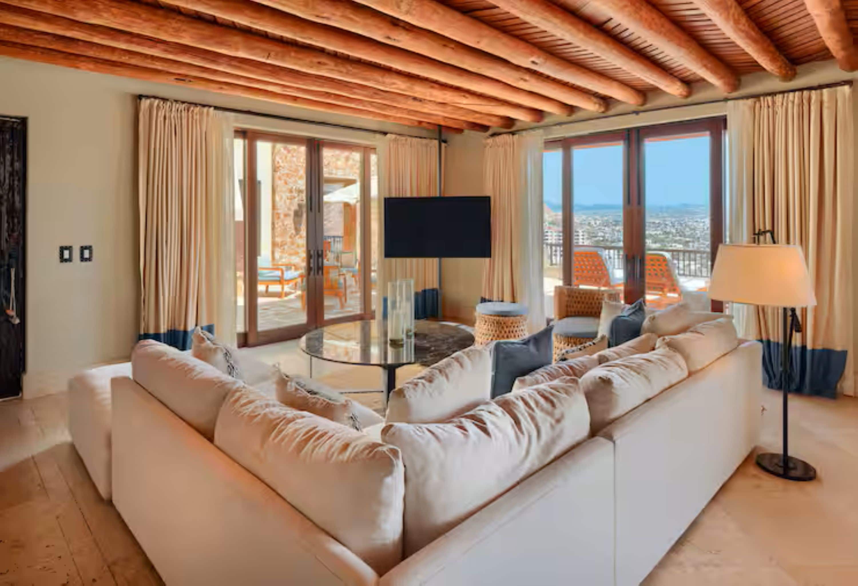 A living room with a sofa, a glass coffee table, and a flat-screen TV. There are large windows with curtains, a view of the city, and a wooden ceiling.