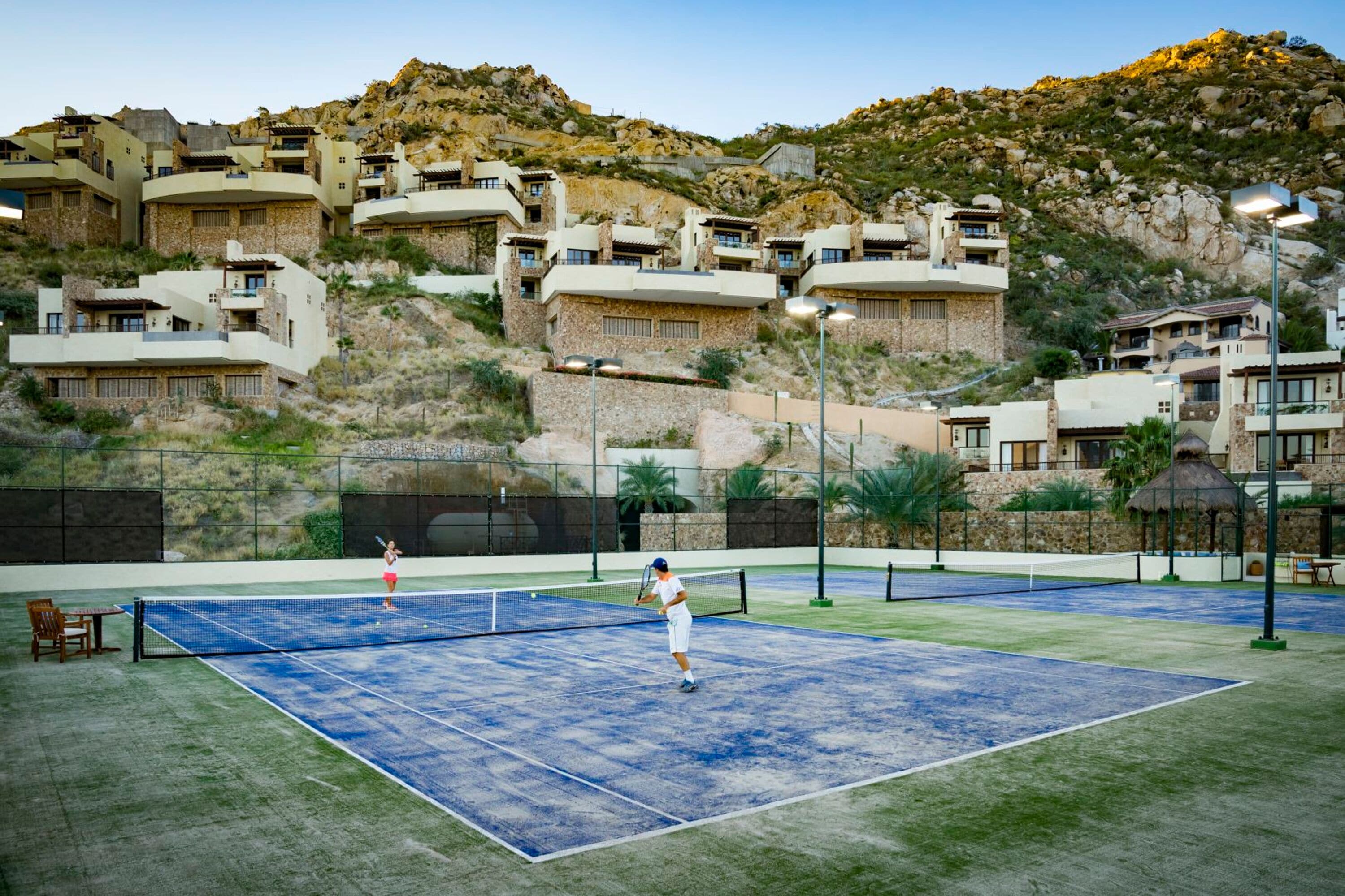 A tennis court with two players, surrounded by hillside buildings.