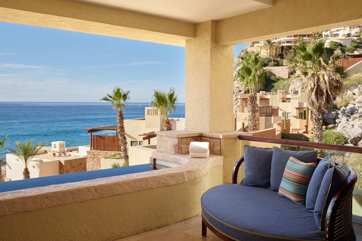 Ein Balkon mit Blick auf das Meer, ein Stuhl mit Kissen und ein kleiner Tisch mit einem weißen Gegenstand.