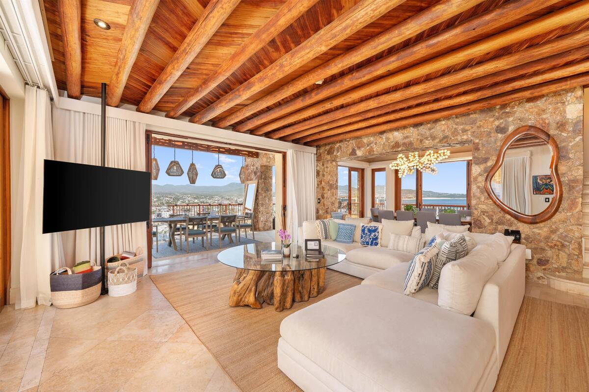 Amplia sala de estar con una gran televisión de pantalla plana, una mesa de centro redonda y un sofá confortable. La habitación cuenta con vigas de madera a la vista, paredes de piedra y grandes ventanales con vistas al paisaje.