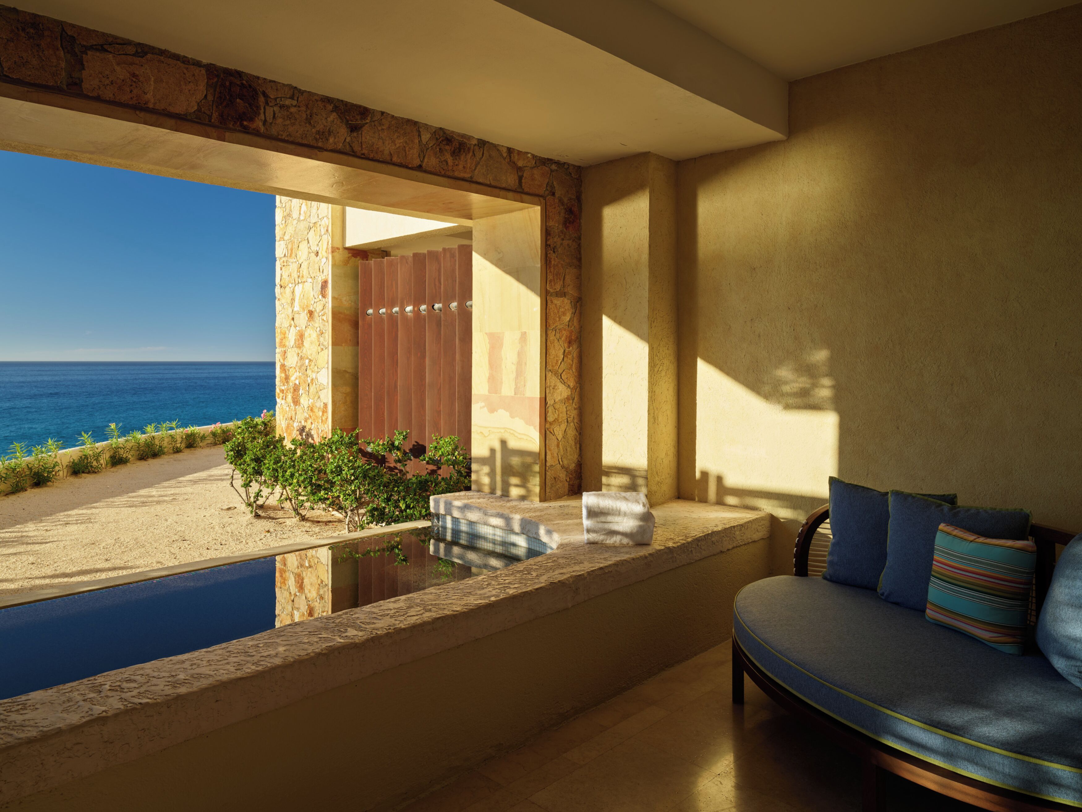 A room with a view of the sea, a pool, and a seating area with cushions.