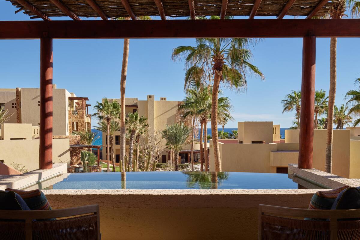 Zona de piscina con vista despejada al océano, rodeada de palmeras y una pérgola de madera.