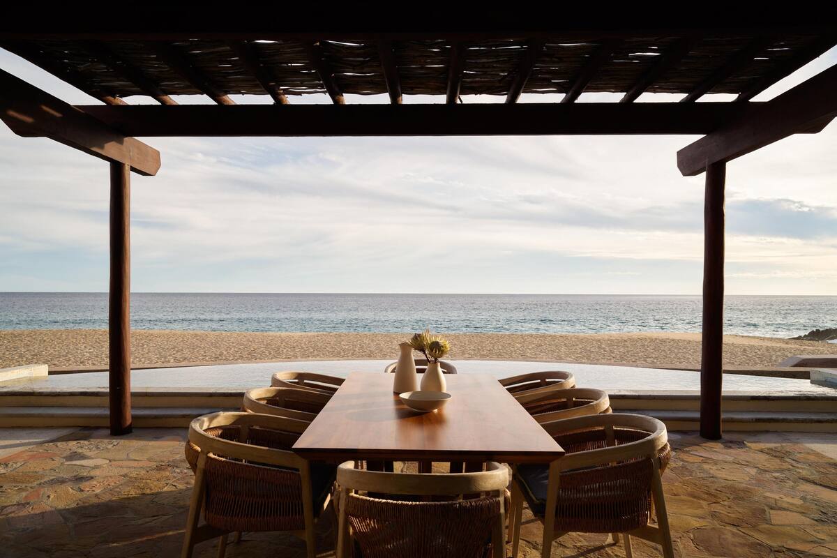 Una mesa de madera con sillas de mimbre bajo una pérgola, con vista a una playa y al mar.