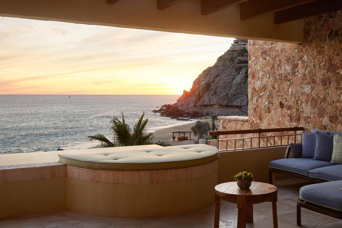 Un balcón con vista al mar y un acantilado, una bañera de hidromasaje circular, una mesa de madera y un sofá azul.