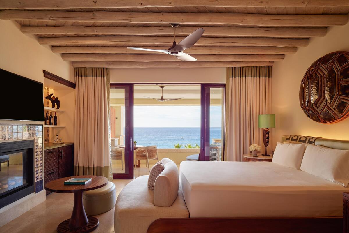 Habitación de hotel con una cama grande, televisión, chimenea, balcón con vistas al mar y ventilador de techo.