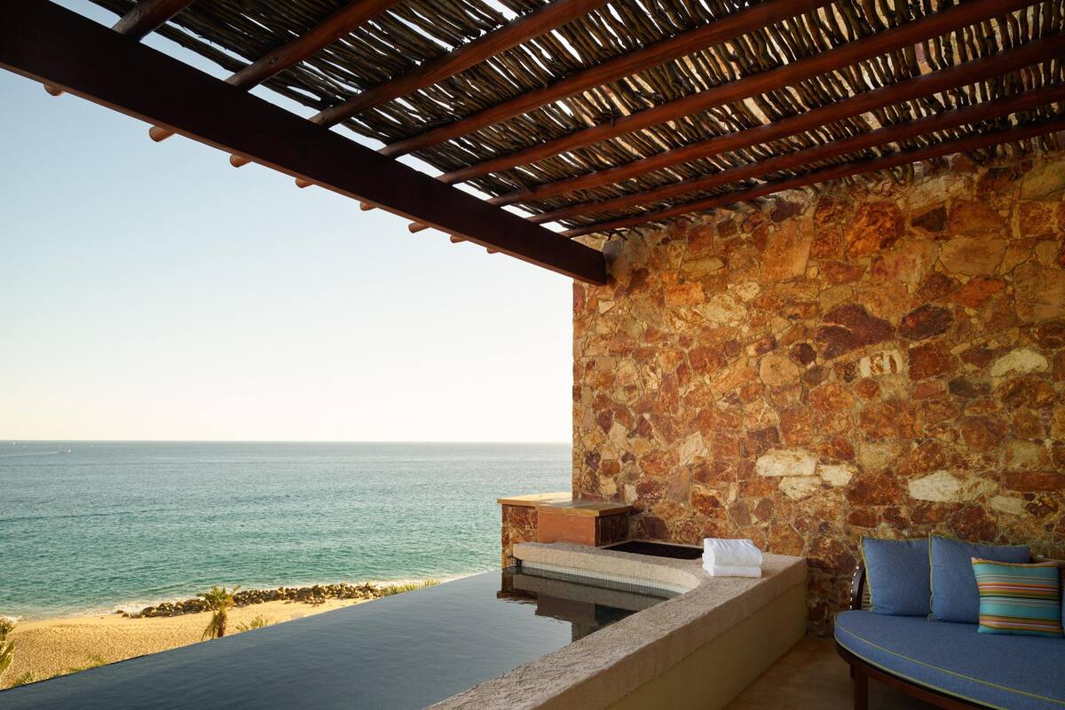 Una piscina infinita de paredes de piedra con una pérgola de madera sobre ella, con vistas a una playa y al mar.