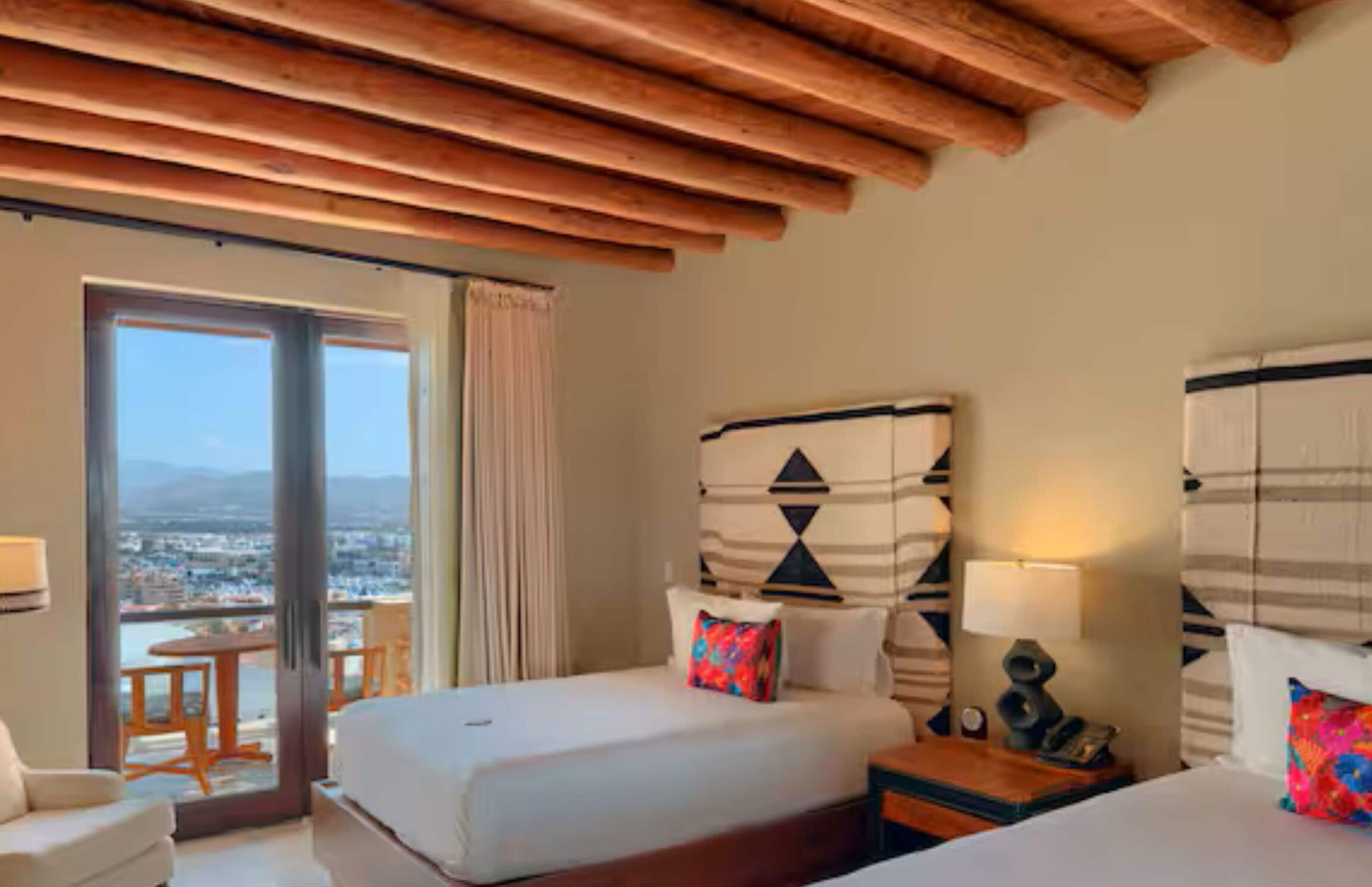 A hotel room with a large bed, a small table, a chair, and a view of the city through a balcony.