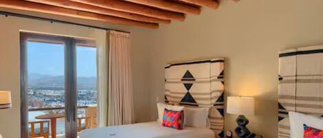 A hotel room with a large bed, a small table, a chair, and a view of the city through a balcony.