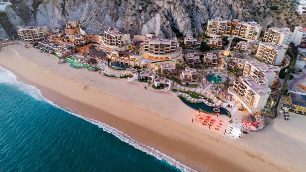 Vista aérea de un complejo turístico costero con varias construcciones, una gran piscina y una playa.