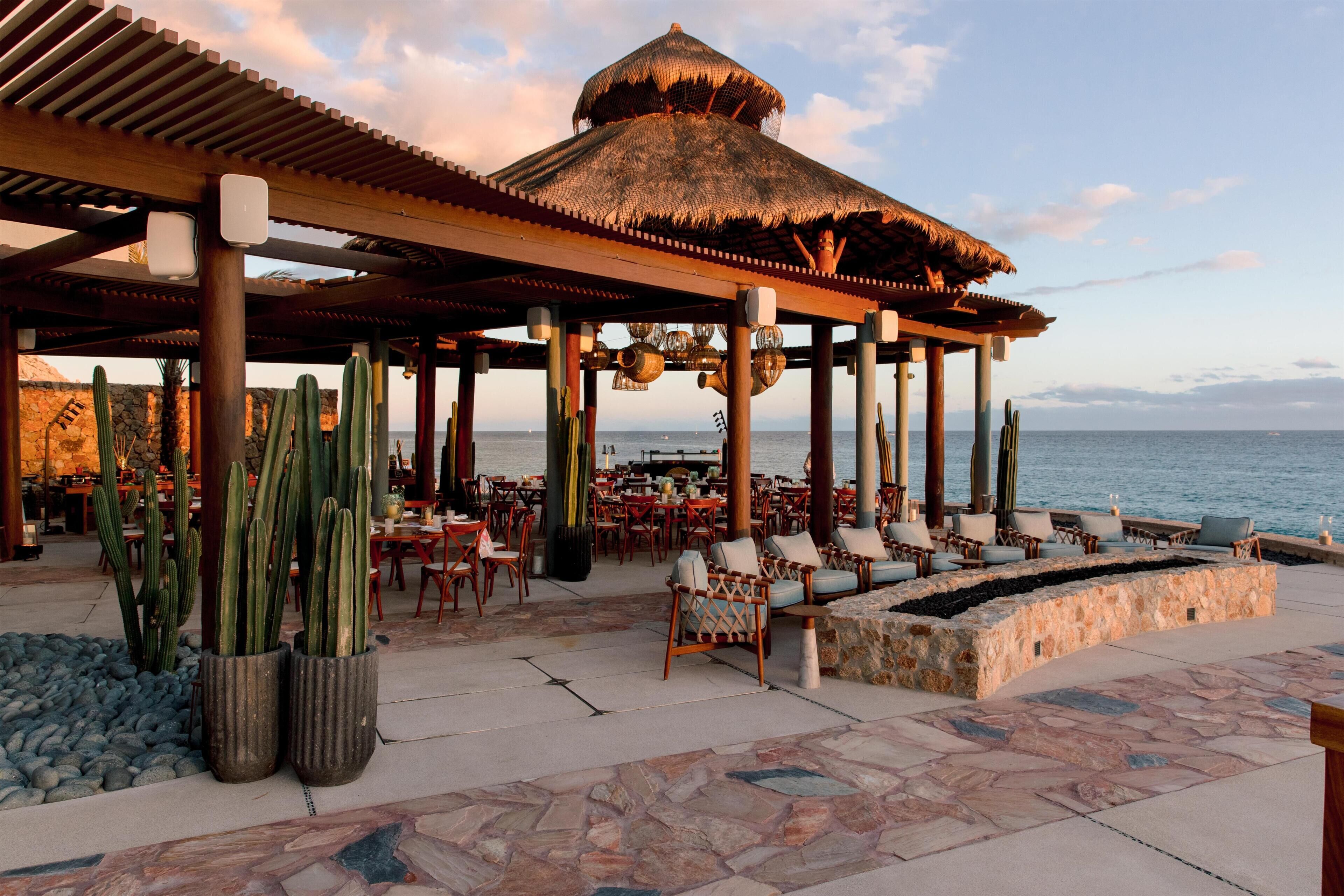 A covered outdoor seating area with tables and chairs, a thatched-roof structure, and cacti.