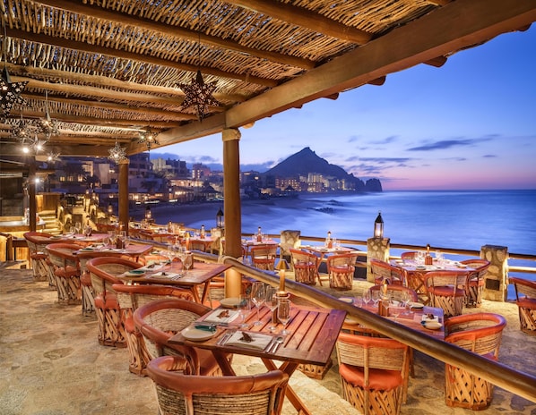 Un restaurante al aire libre con mobiliario de mimbre y vistas al océano y una montaña al atardecer.