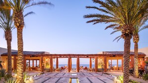 Un hotel frente a la playa con un área de estar al aire libre cubierta, palmeras y un patio empedrado.