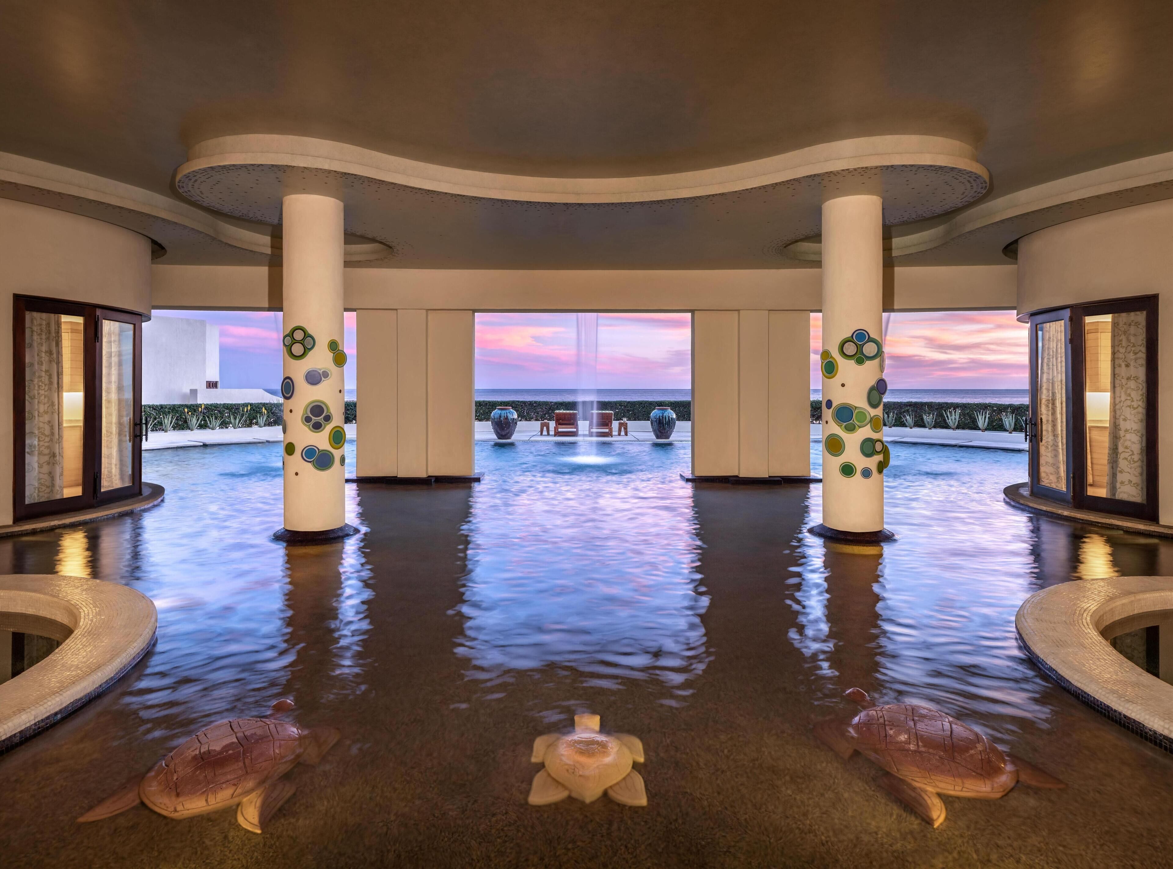A luxurious indoor pool area with a view of the ocean at sunset, featuring two large columns with decorative patterns, a central water feature, and two turtle sculptures on the floor.