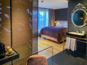 A hotel room with a large bed, a bathroom with a mirror and sink, and a glass partition with a screen. - La Mansion Vi Vadi Hotel (Munich)
