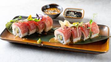 A plate of sushi with two types of rolls, garnished with green leaves and sesame seeds, and accompanied by two small bowls of dipping sauce.