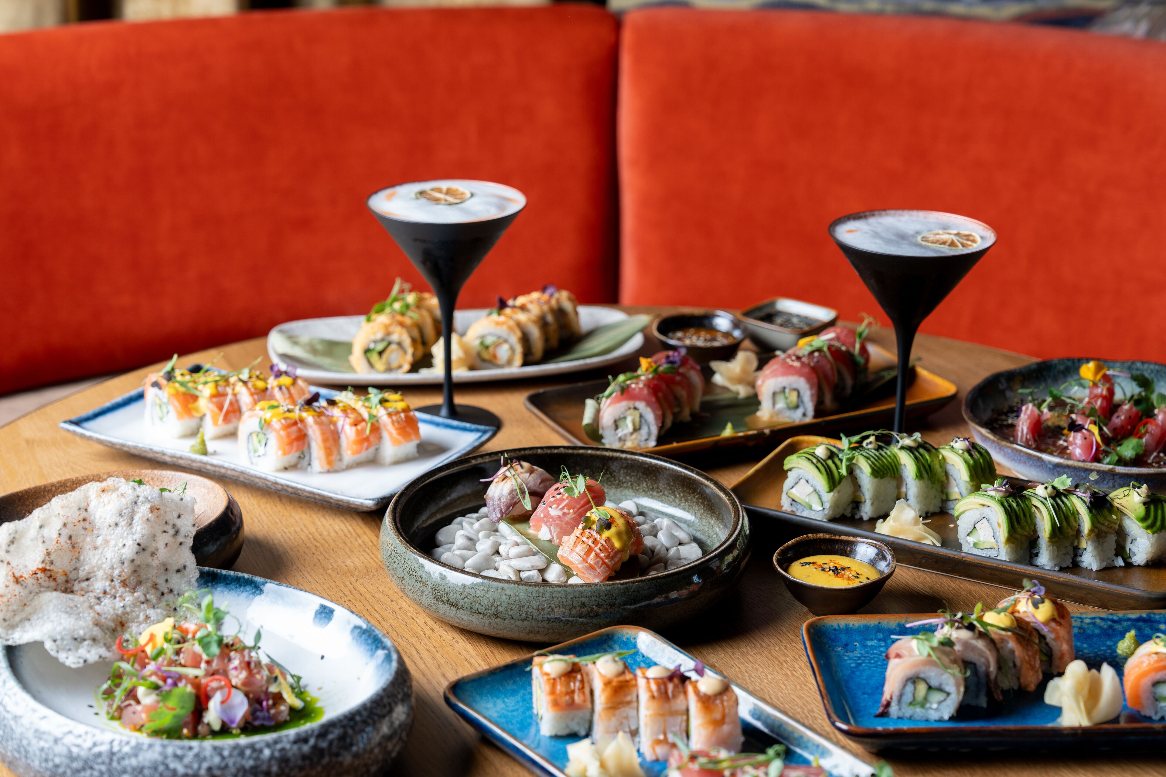 Forskellige sushi retter på et træbord med en rød polstret sofa i baggrunden.