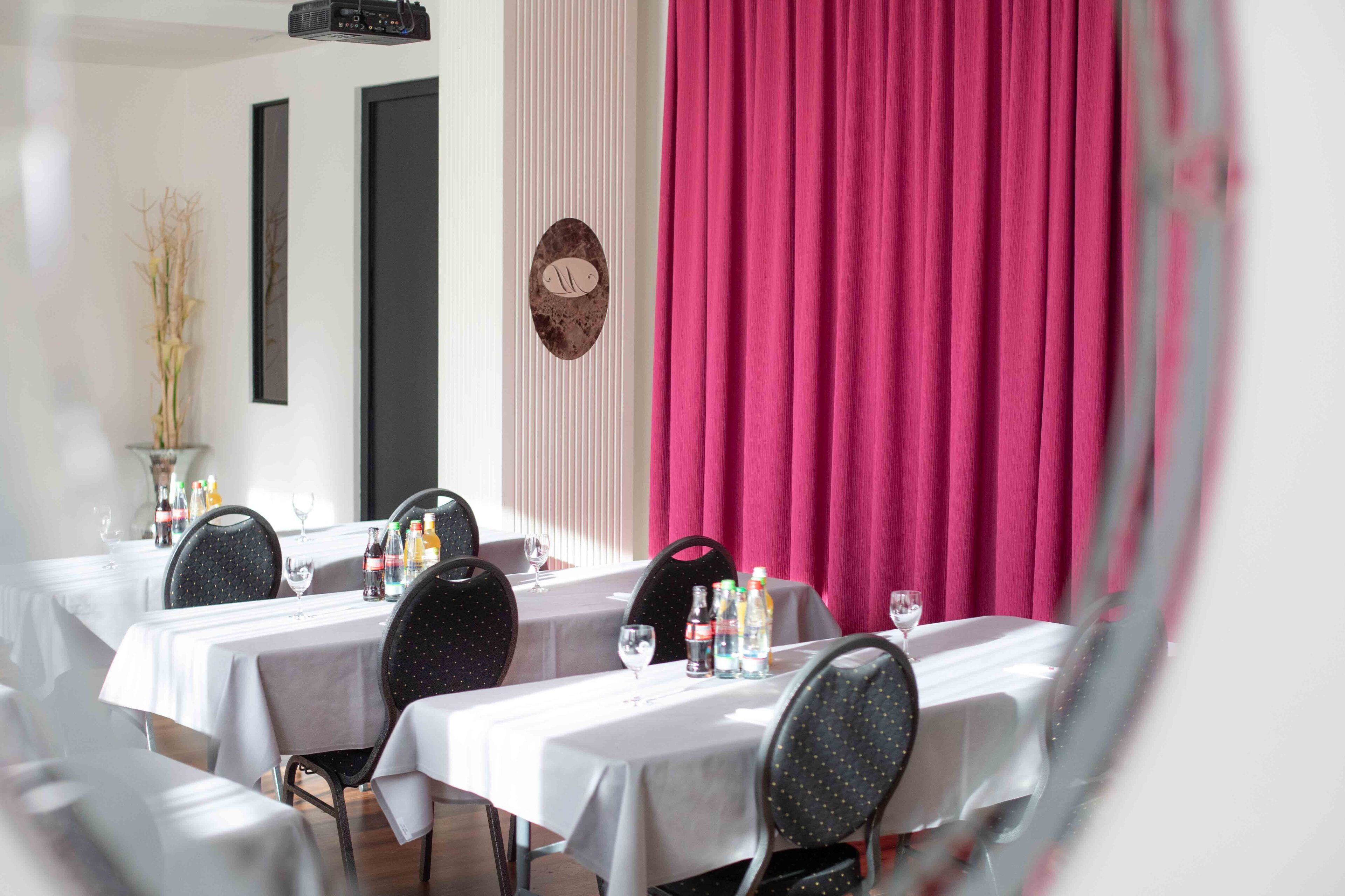 A dining area with tables set for two, each with a white tablecloth and black chairs. There are bottles and glasses on the tables, and a vase with branches on one side.