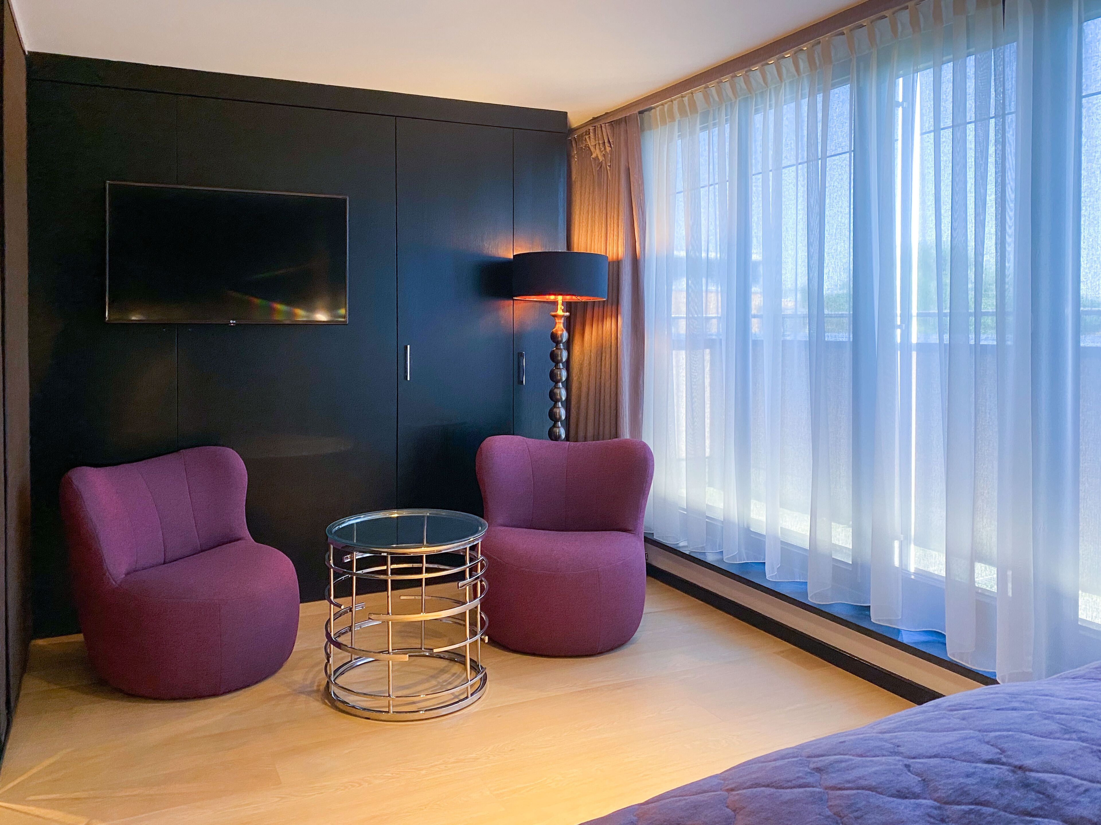 A modern hotel room with a large bed, two armchairs, a small round table, a TV mounted on the wall, and a large window with sheer curtains.