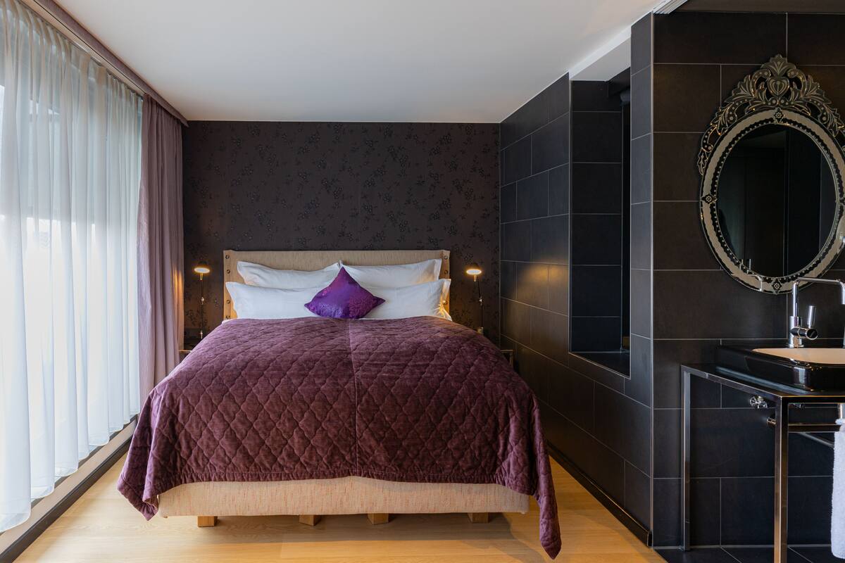 A modern bedroom with a large bed, a dark wall with a mirror, and a window with sheer curtains.