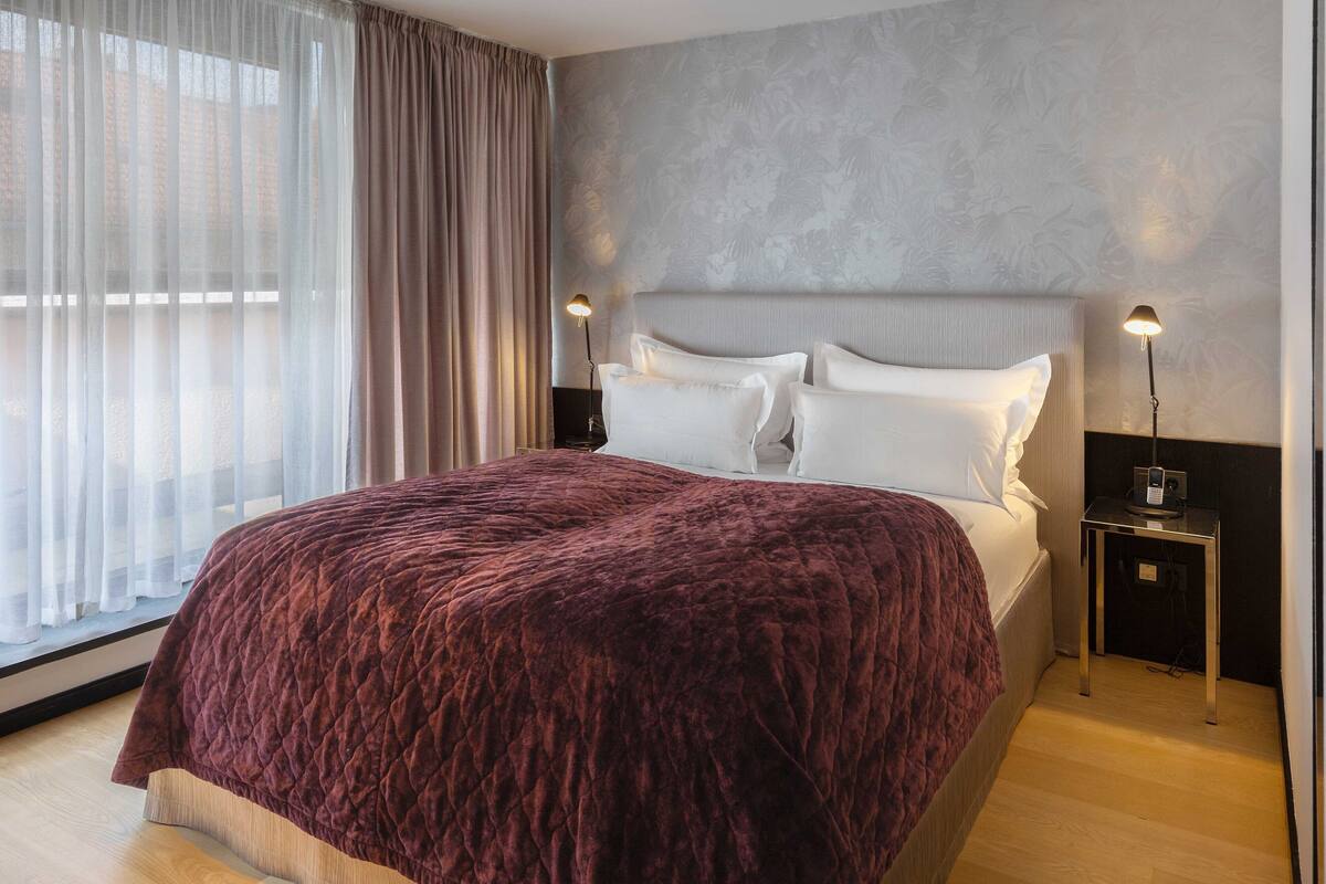 A modern hotel room with a large bed, a nightstand, a lamp, and a view of the outdoors through sheer curtains.