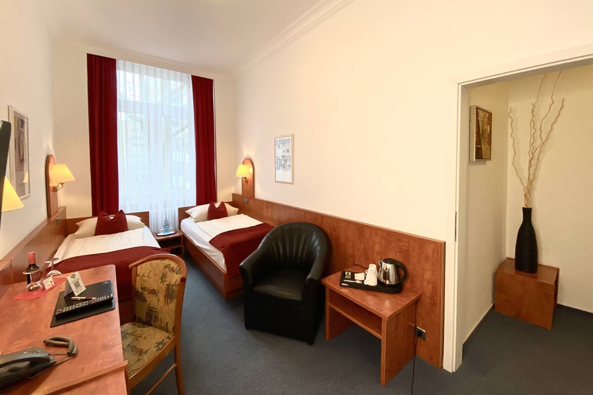 A hotel room with two beds, a desk with a telephone, a chair, a small table with a kettle, and a vase with branches.