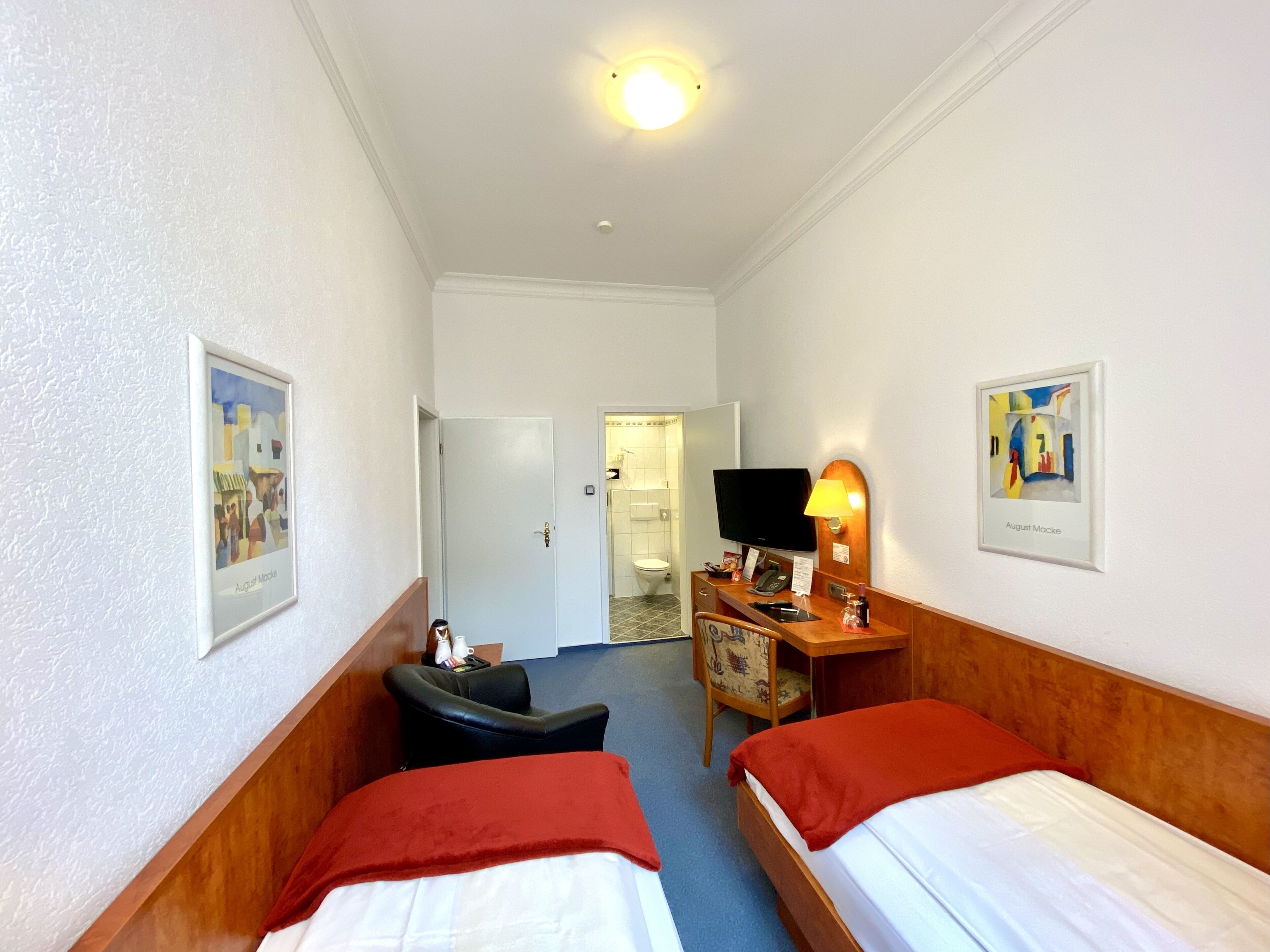 A hotel room with two single beds, a desk with a television, a chair, and a small table with a lamp.