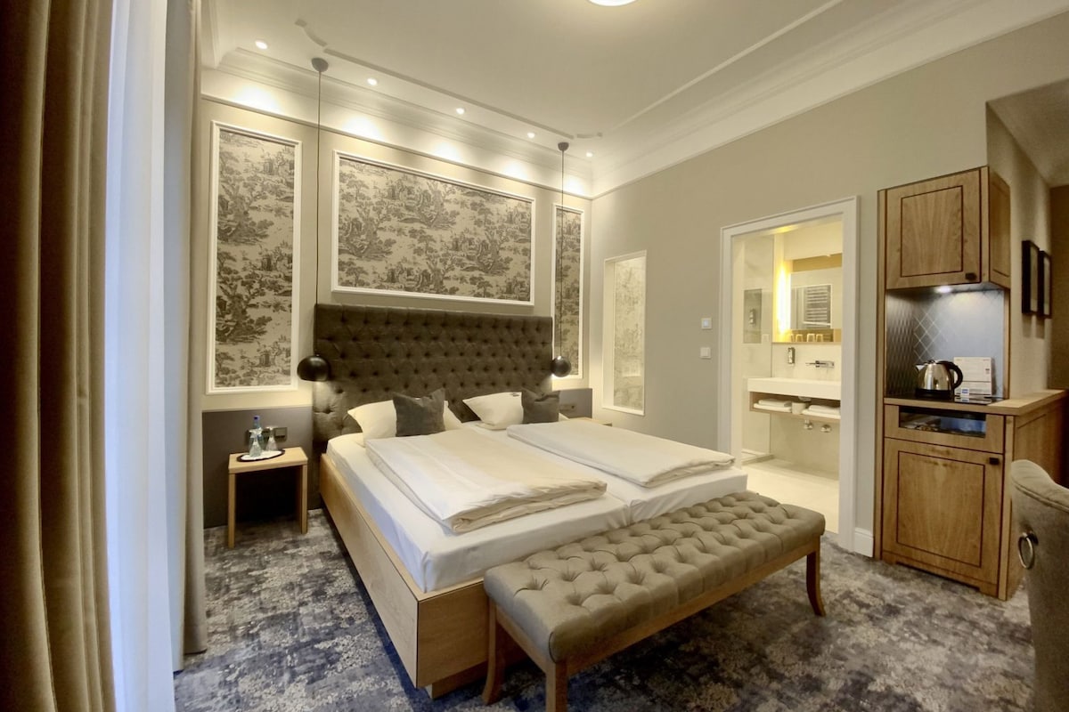 A modern hotel room with a large bed, a bench, a nightstand, a mirror, and a kitchenette.