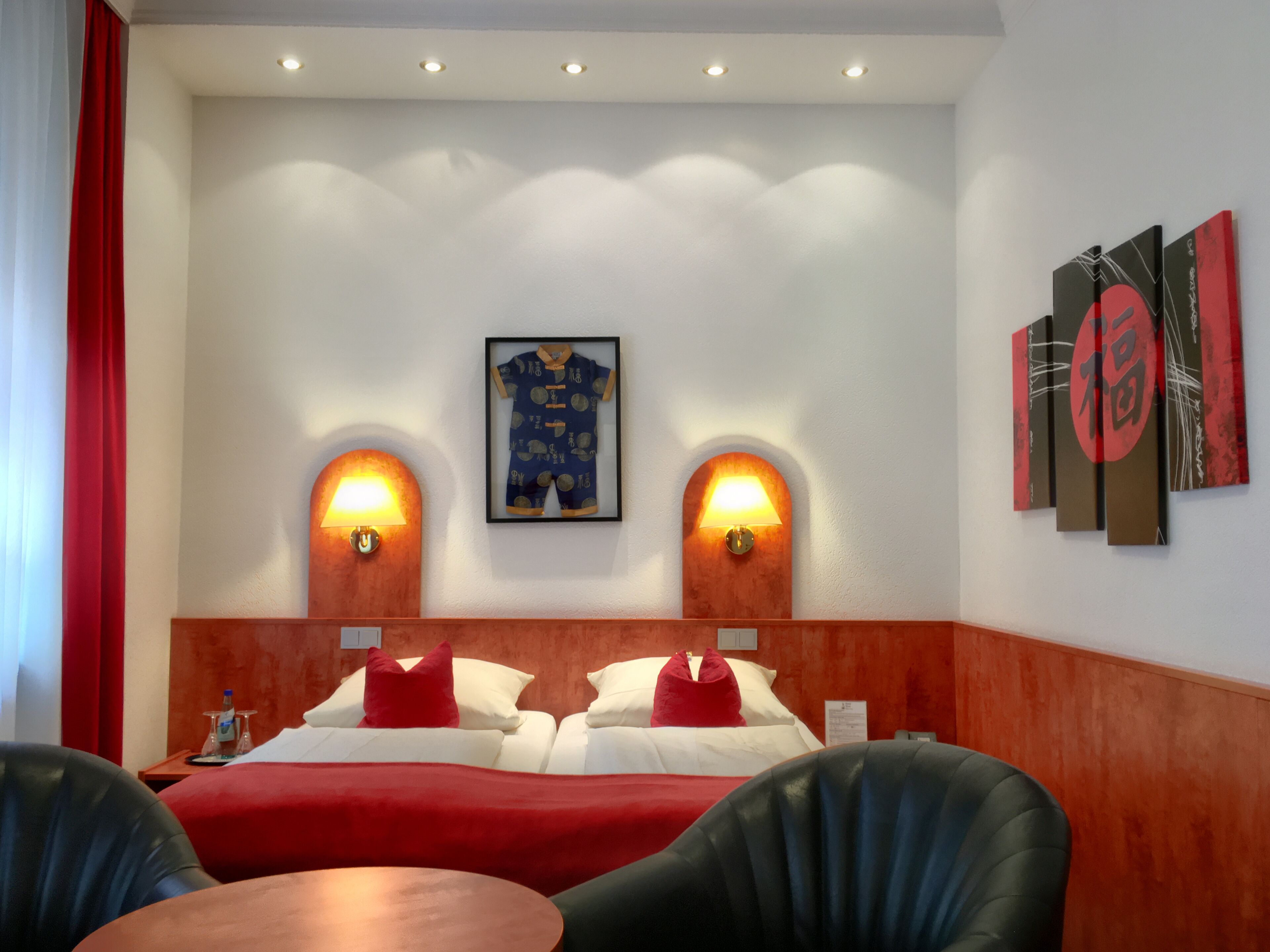 Habitació d'hotel amb un llit, dues làmpades penjades a la paret, un quadre amb marc i una cortina vermella.