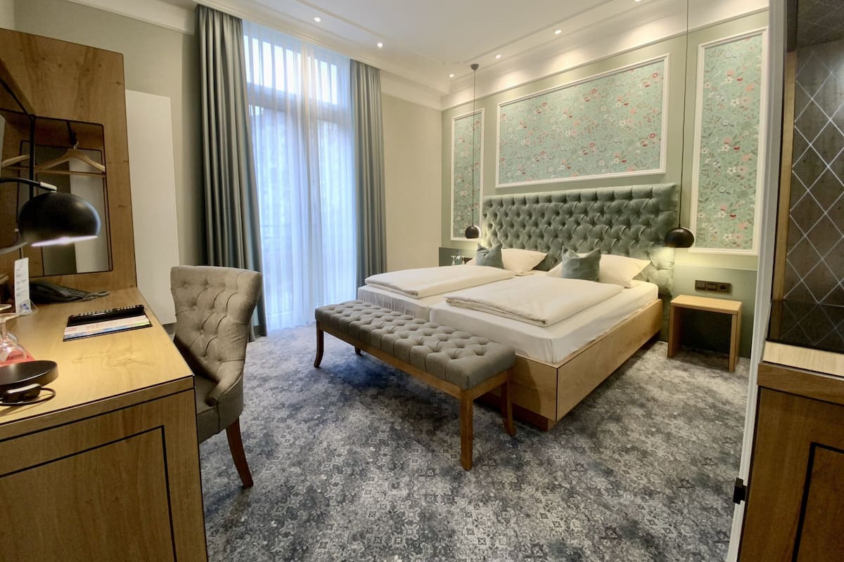 A hotel room with a large bed, two bedside tables, a desk with a chair, a small bedside table, and a large window with curtains.