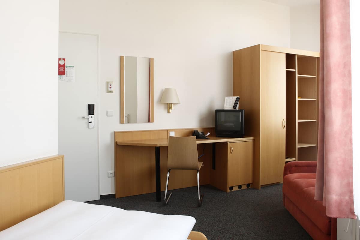 Ein Hotelzimmer mit einem Bett, einem Schreibtisch mit Computer und Fernseher, einem Sessel, einem Kleiderschrank und einem roten Vorhang.