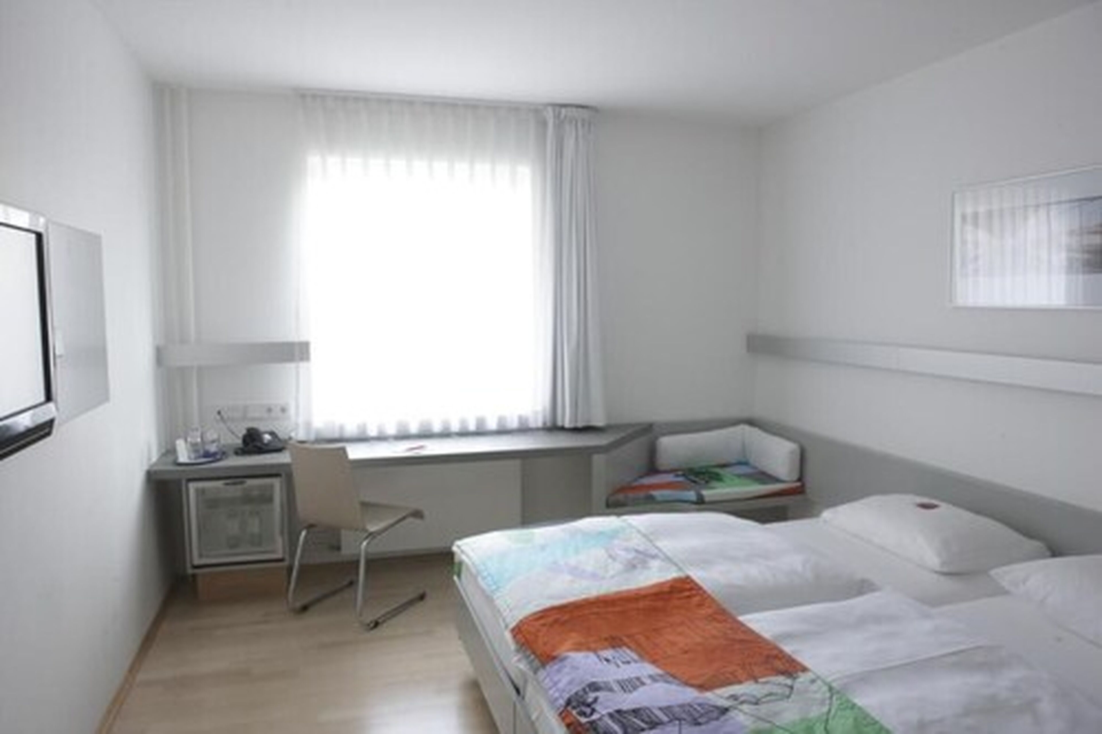 Ein Hotelzimmer mit Bett, Schreibtisch, Stuhl, einem kleinen Schrank und einem Fenster mit Vorhängen.