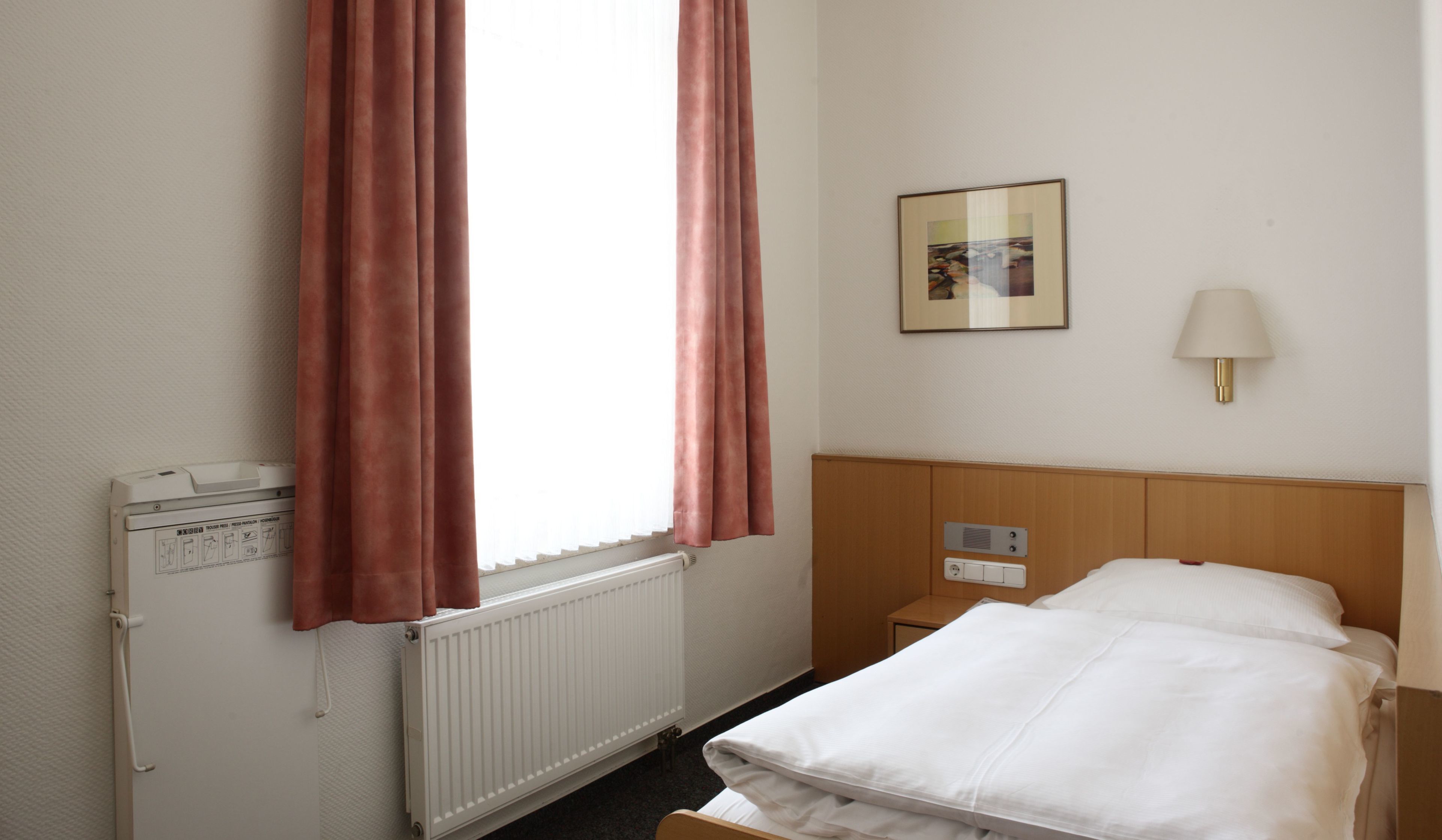 Une chambre d’hôtel avec un lit, une table de chevet, une lampe, un radiateur et des rideaux.