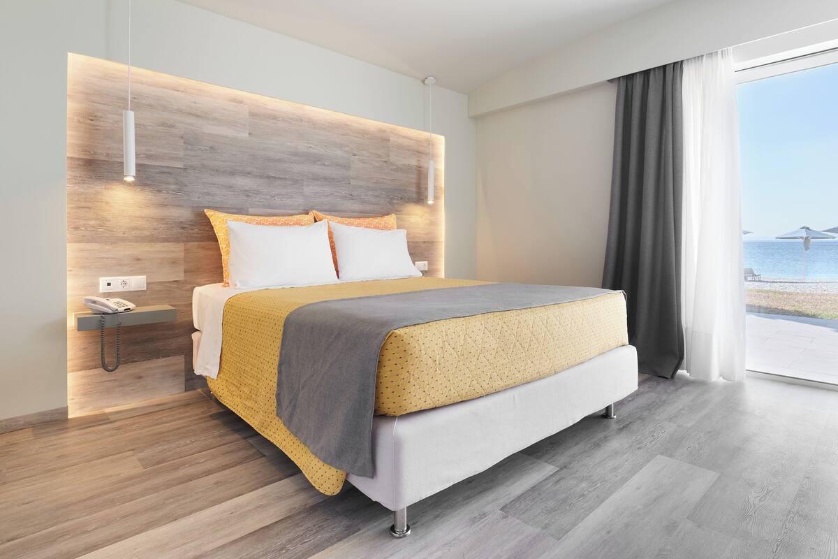 Quarto de hotel moderno com cabeceira de madeira, cama com edredom amarelo e cinzento, telefone de apoio e vista para o mar através de grandes janelas.