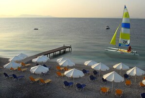 A beach with rows of umbrellas and lounge chairs, a sailboat with a yellow and blue sail, and a pier extending into the water.