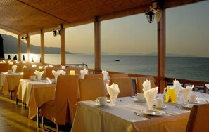 A restaurant with tables set for dining, overlooking a body of water with mountains in the background.