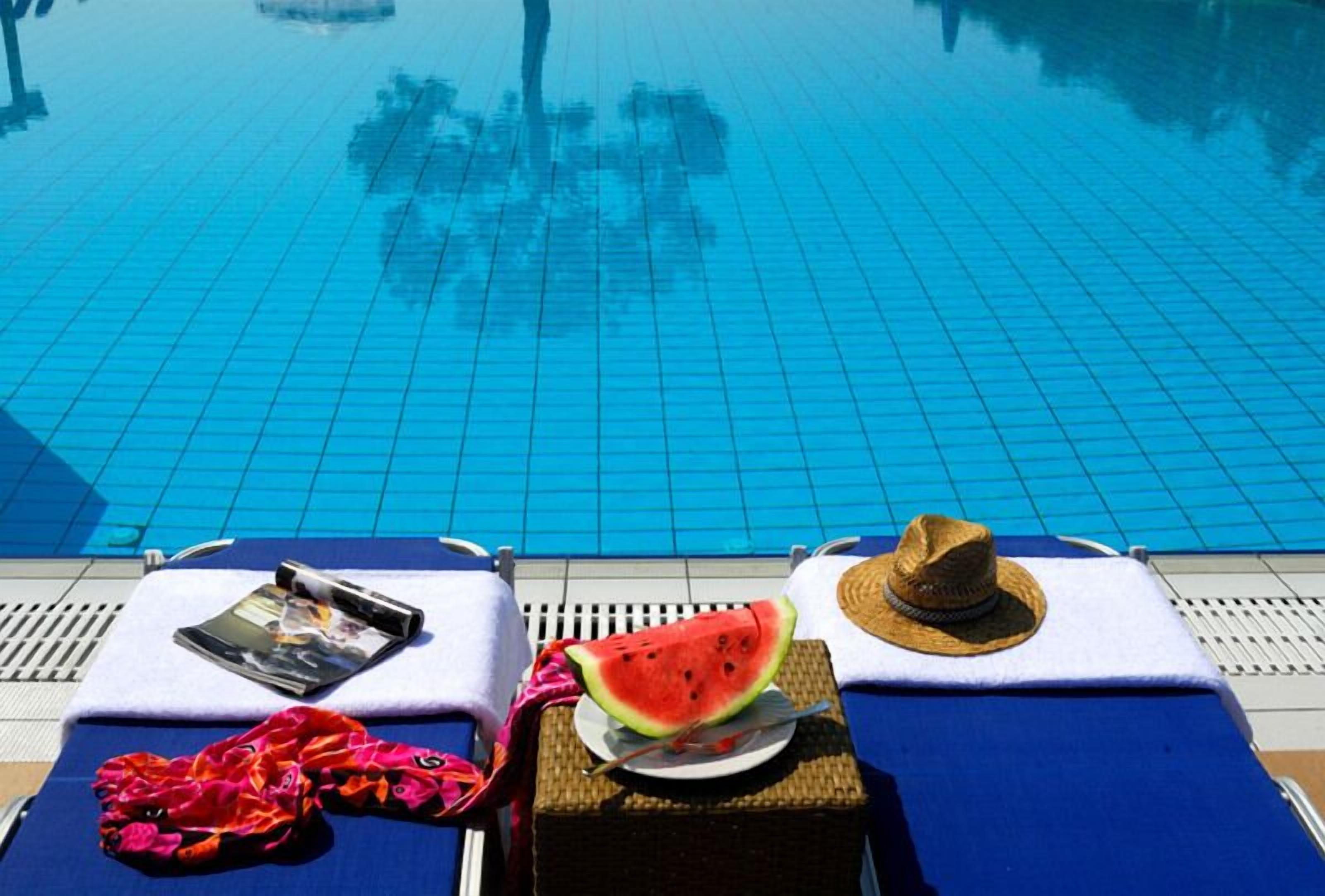 Un área de descanso junto a la piscina con suelo de baldosas azules, un sombrero, una tajada de sandía y una revista sobre una toalla.