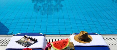 Un área de descanso junto a la piscina con suelo de baldosas azules, un sombrero, una tajada de sandía y una revista sobre una toalla.