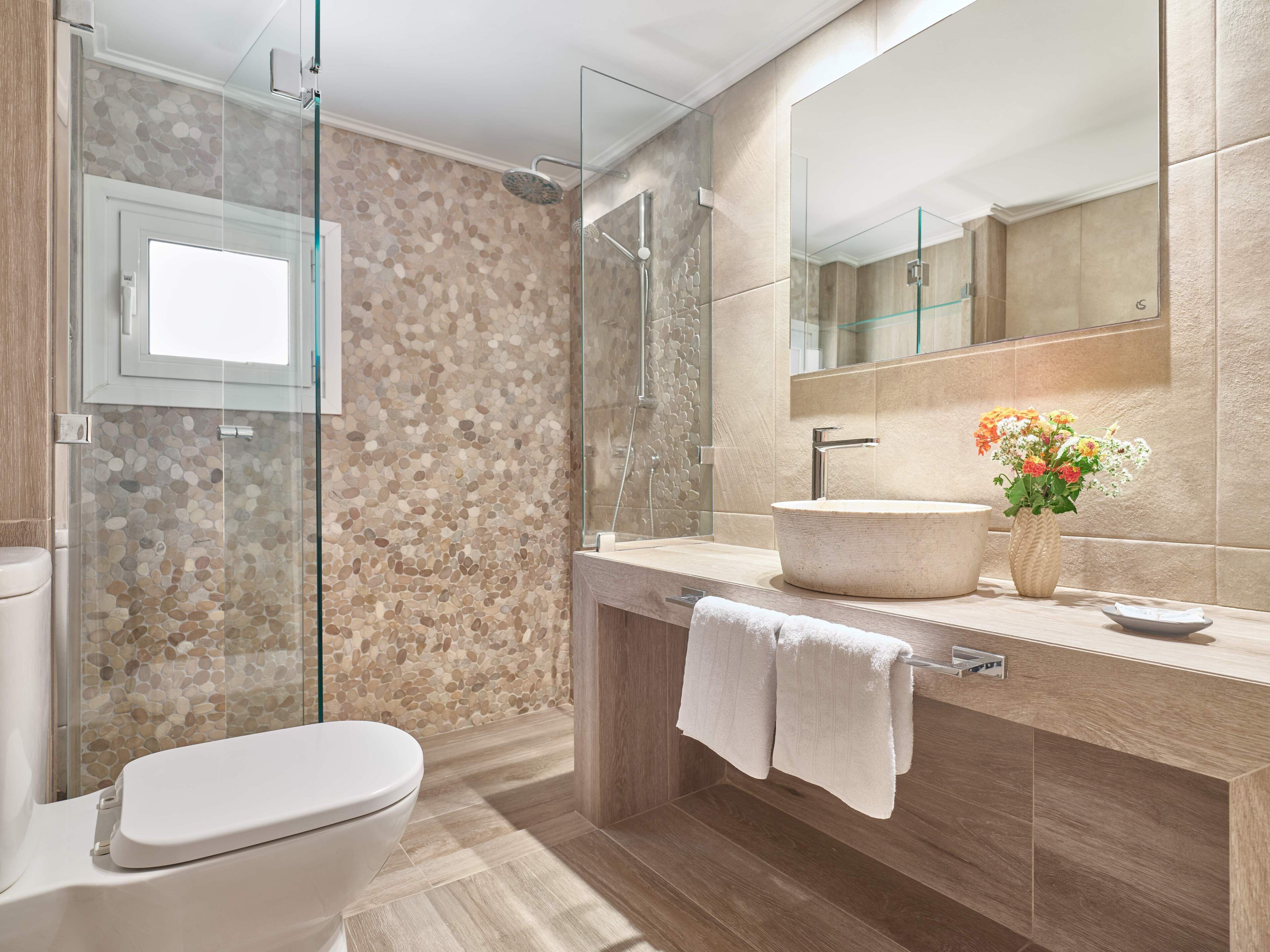 A modern bathroom with a glass-enclosed shower, a freestanding toilet, a vanity with a large mirror, and a vase with flowers.