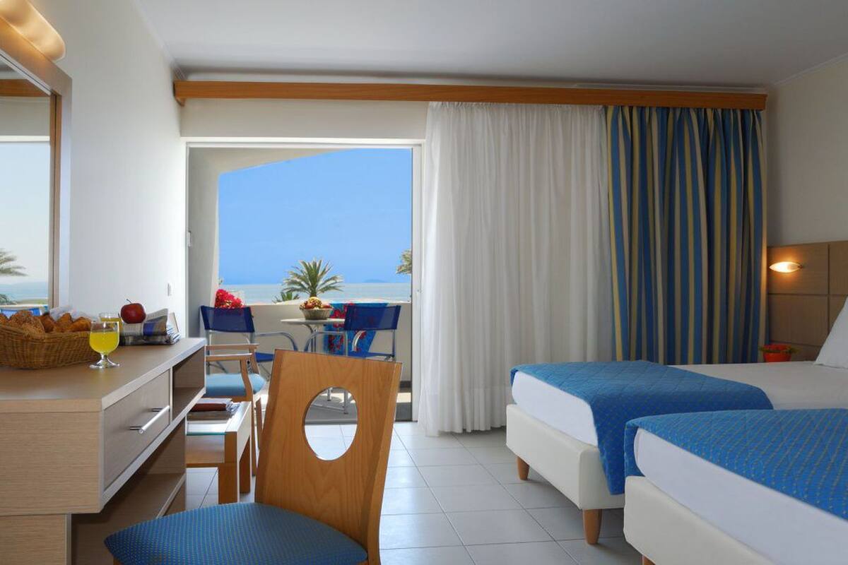 Quarto de hotel com duas camas, uma escrivaninha com cadeira, zona de refeições com mesa e cadeiras, e vista para o mar.