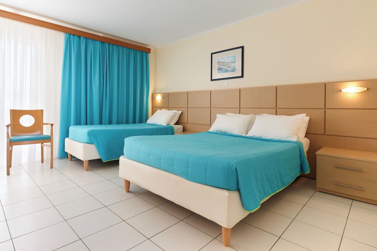 Quarto de hotel com duas camas, uma cadeira de madeira, uma mesa de cabeceira e um quadro na parede.