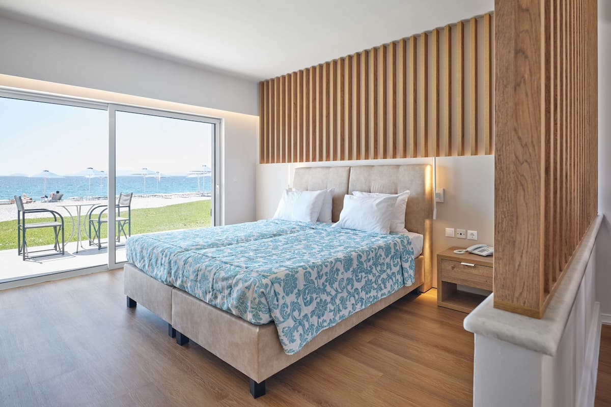 Quarto moderno com uma cama grande, cabeceira de madeira, mesa de cabeceira e vista para a praia por uma porta de correr de vidro.