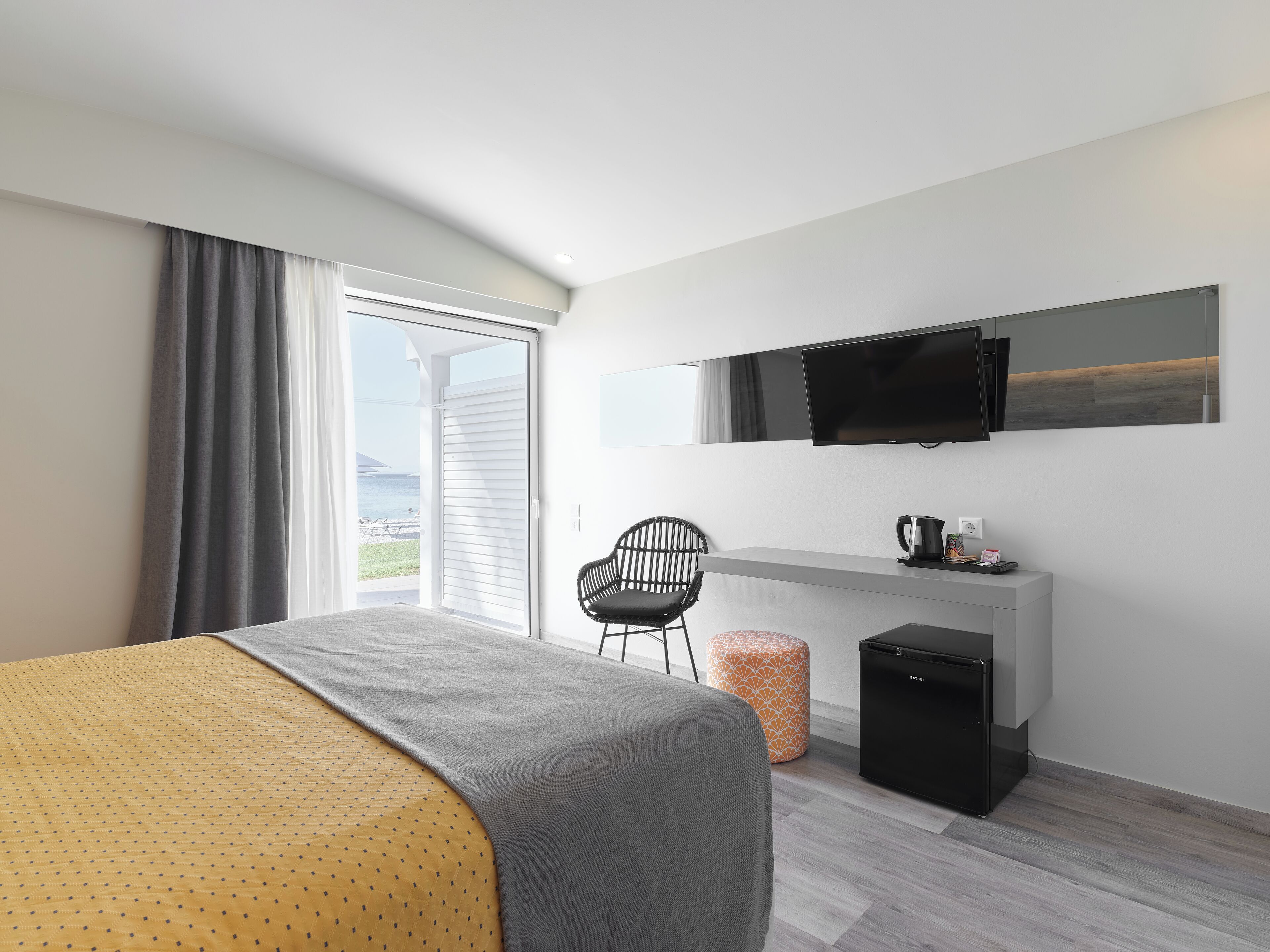 A modern hotel room with a bed, a desk, a chair, a television, and a view of the sea.