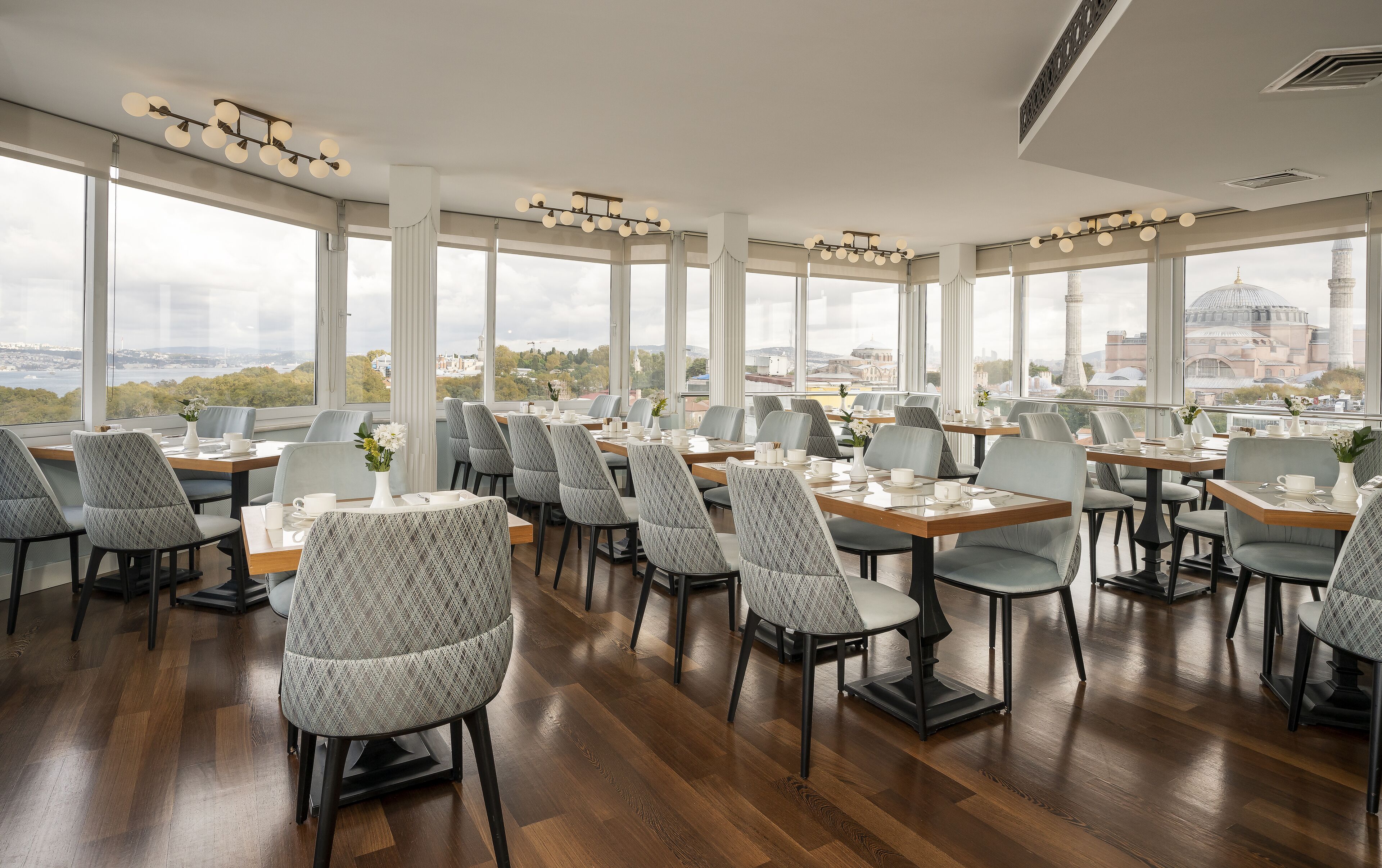Un restaurante con varias mesas y sillas, amplias ventanas que ofrecen vistas a la ciudad, e iluminación moderna.