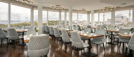 Un restaurante con varias mesas y sillas, amplias ventanas que ofrecen vistas a la ciudad, e iluminación moderna.