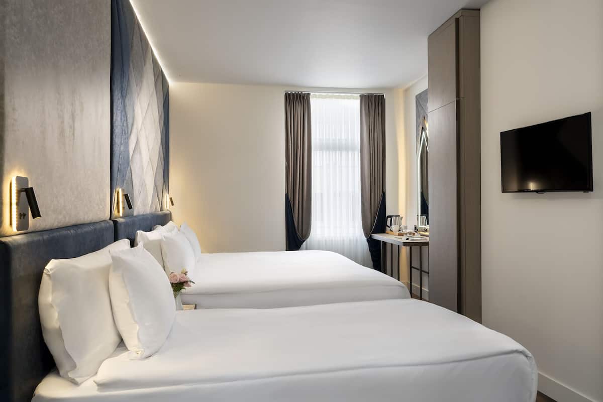 Ein Hotelzimmer mit zwei Betten, einem Flachbildfernseher, einem Schreibtisch mit Kaffeemaschine und einem großen Fenster mit Vorhängen.