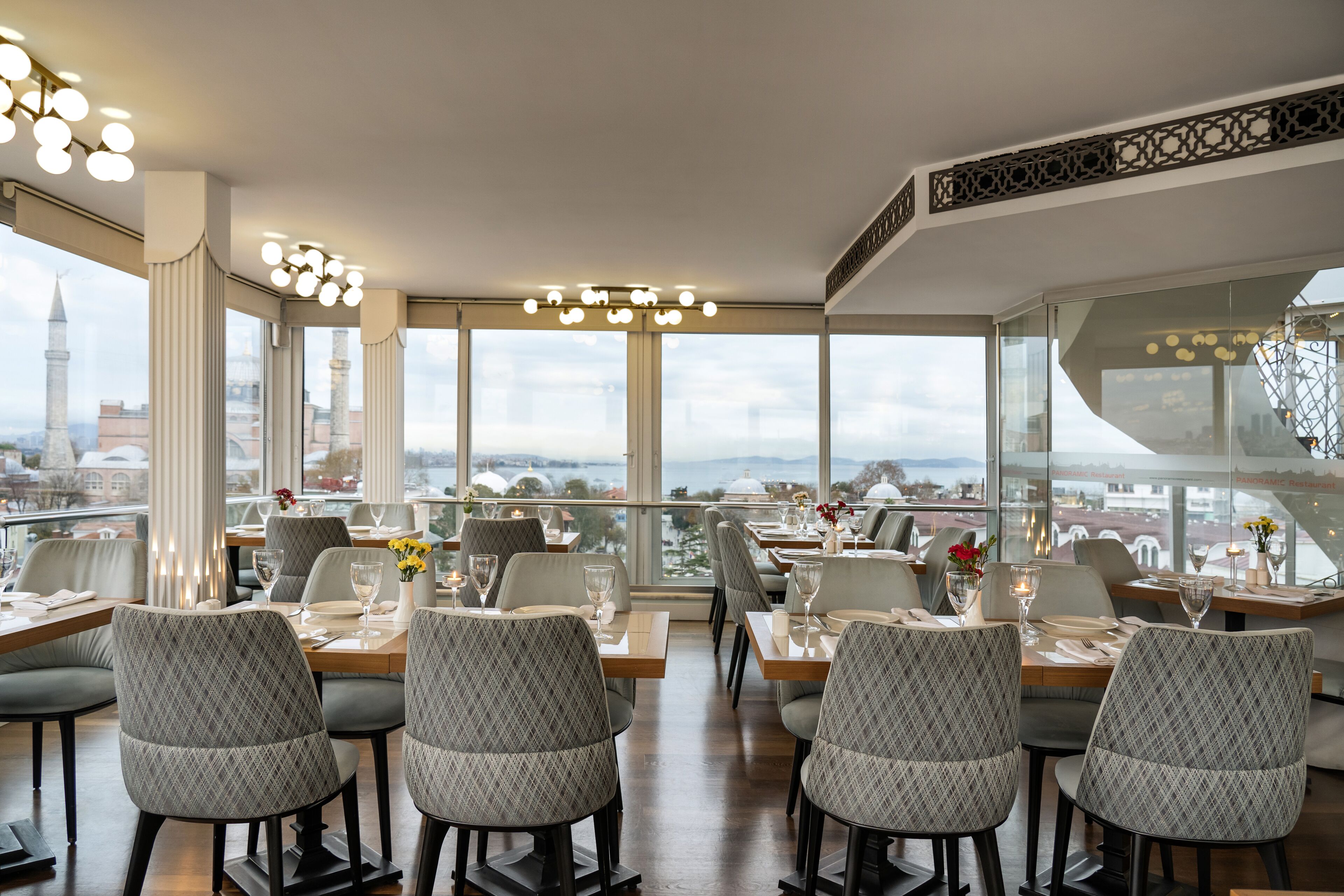 A restaurant with a view of a cityscape, featuring tables set with wine glasses and flowers.
