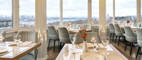 A restaurant with a table set for dining, large windows, and a city view.